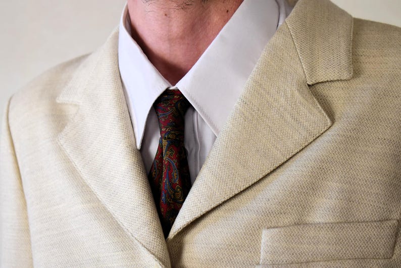 Pu&ograve; includere: Un blazer beige in lino con una camicia bianca con colletto e una cravatta con motivo paisley. La cravatta &egrave; rossa, blu e gialla.