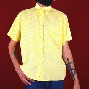 Puede incluir: Camisa amarilla de manga corta con estampado de lunares blancos. Se combina con vaqueros azules. El fondo es de color rojo.