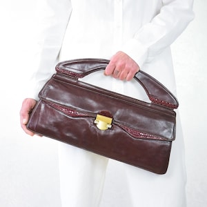 Peut inclure: Sac à main bordeaux vintage avec fermoir et poignée dorés. Le sac présente un accent texturé brun rougeâtre le long du bord supérieur. Le sac rectangulaire est tenu par une personne vêtue de blanc.