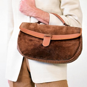 Pode incluir: Uma mala de camurça castanha com alça e fecho de couro. A mala tem formato curvo e uma borda texturizada. A mala está sendo segurada por uma pessoa vestindo um blazer creme e calças de veludo cotelê marrons.