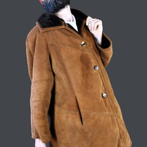 May include: A brown suede shearling coat with a black fur collar and three gold buttons. The coat is worn by a person wearing a red beanie and sunglasses.