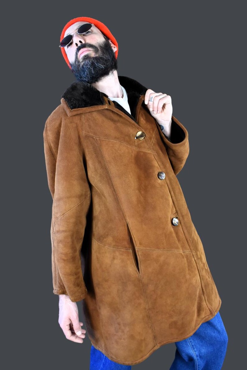 May include: A brown suede shearling coat with a black fur collar and three gold buttons. The coat is worn by a person wearing a red beanie and sunglasses.