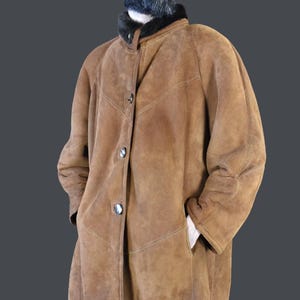 May include: A brown suede shearling coat with a black fur collar and button closure. The coat is worn by a person wearing a red beanie and sunglasses.