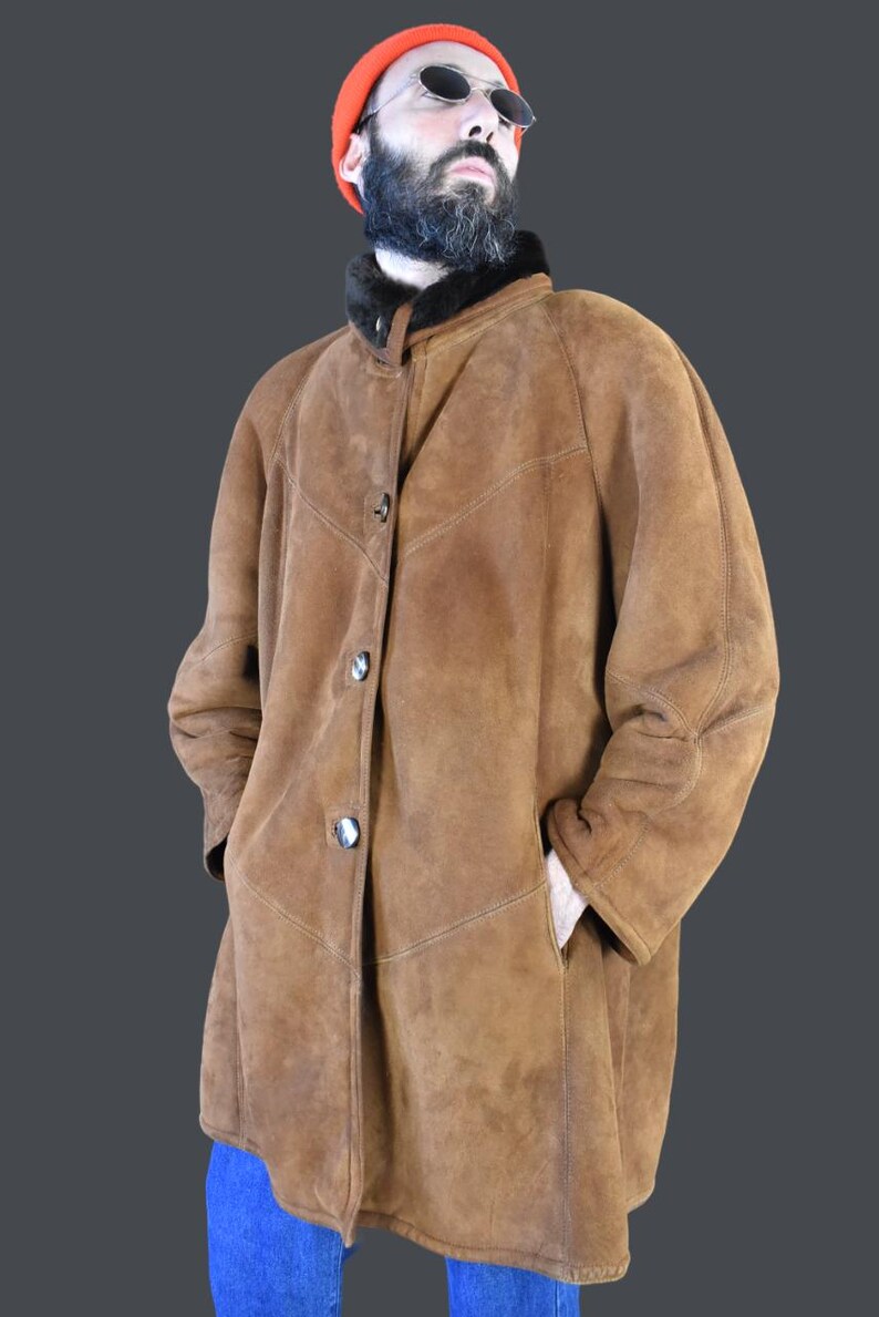 May include: A brown suede shearling coat with a black fur collar and button closure. The coat is worn by a person wearing a red beanie and sunglasses.