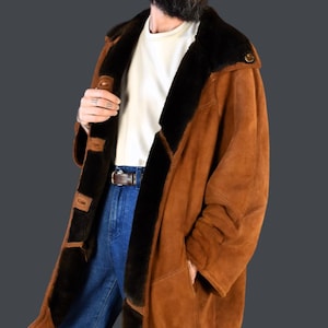May include: A brown suede shearling coat with a black fur collar and lining. The coat has a button closure and two pockets. The model is wearing blue jeans and a white t-shirt.