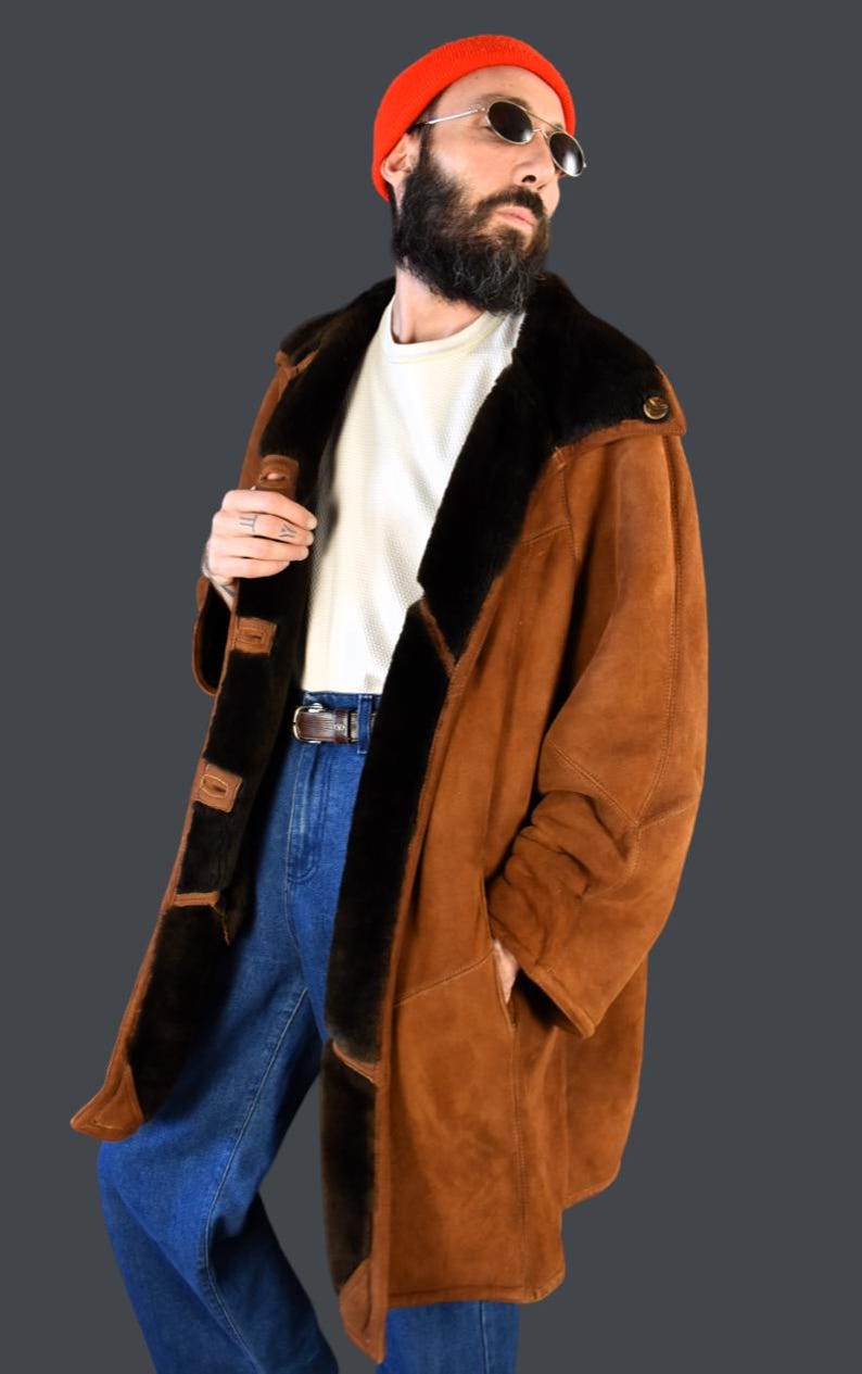 May include: A brown suede shearling coat with a black fur collar and lining. The coat has a button closure and two pockets. The model is wearing blue jeans and a white t-shirt.