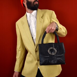 May include: A black leather handbag with a silver circular clasp. The bag is held by a person wearing a yellow plaid blazer, white shirt, black pants, and a red beanie. The background is a solid red.