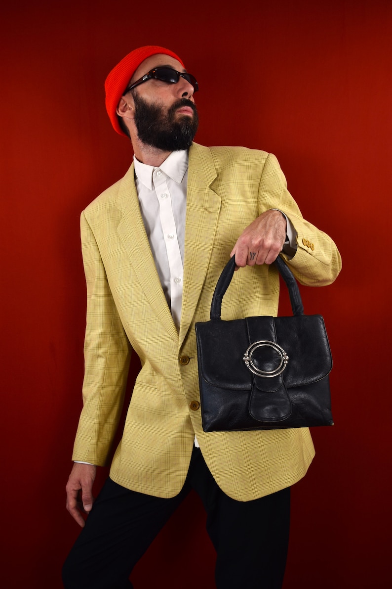 May include: A black leather handbag with a silver circular clasp. The bag is held by a person wearing a yellow plaid blazer, white shirt, black pants, and a red beanie. The background is a solid red.