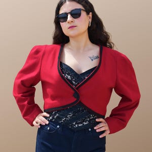 May include: A red cropped jacket with black trim and a single button closure. The jacket has a slightly puffed shoulder and is worn over a black lace top.