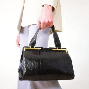 Peut inclure: Un sac à main noir avec un motif peau de serpent et des détails dorés. Le sac est de forme rectangulaire avec une structure rigide et une poignée supérieure. Il est porté par une personne vêtue d'un blazer crème et d'un pantalon gris clair.