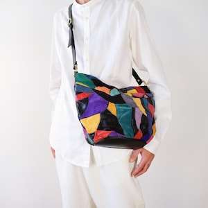 May include: A crossbody bag featuring a patchwork design of multi-coloured leather pieces in shades of black, red, yellow, purple, teal, and orange. The bag includes a black leather shoulder strap with gold-toned metal hardware and a solid black base.