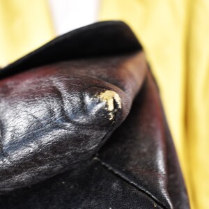 May include: Close-up of a black leather bag with visible wear and tear. The leather has a textured appearance with some areas showing scuffing and discoloration. The bag's corner displays a patch of worn material.