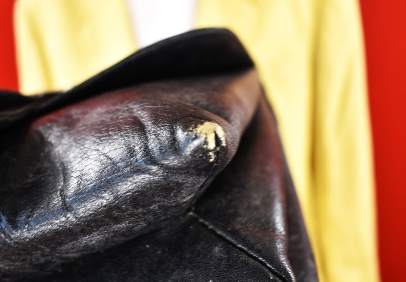 May include: Close-up of a black leather bag with visible wear and tear. The leather has a textured appearance with some areas showing scuffing and discoloration. The bag's corner displays a patch of worn material.
