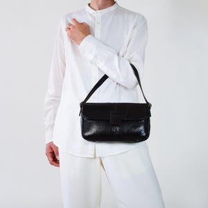 Könnte beinhalten: Schwarze rechteckige Handtasche mit einem strukturierten, reptilienartigen Muster. Die Tasche hat einen Klappenverschluss mit einem rechteckigen Verschluss und einen schwarzen Schulterriemen. Die Tasche wird über der Schulter getragen.