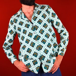 May include: A man wearing a long-sleeved shirt with a geometric pattern in shades of green, white, and yellow. The shirt has a collar and a button-up front. The man is wearing blue jeans and has a beard.