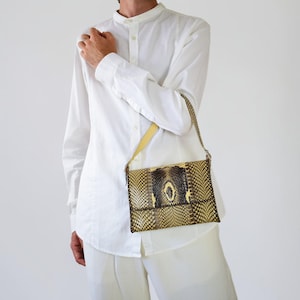 May include: A rectangular handbag featuring a textured snakeskin pattern in shades of yellow, brown, and black. The bag has a slim profile, a fold-over flap closure, and a thin, light yellow shoulder strap, worn against a plain white long-sleeved shirt.