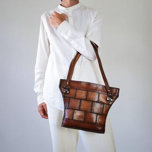 May include: A brown leather tote bag featuring a patchwork design of rectangular panels. The bag has two thin leather shoulder straps attached with metal rings and rivets. The leather has a polished finish with visible stitching along the edges.