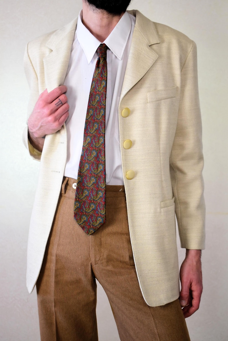 Pu&ograve; includere: Un blazer color crema con una cravatta a fantasia paisley e pantaloni di velluto a coste marroni. Il blazer ha tre bottoni dorati.