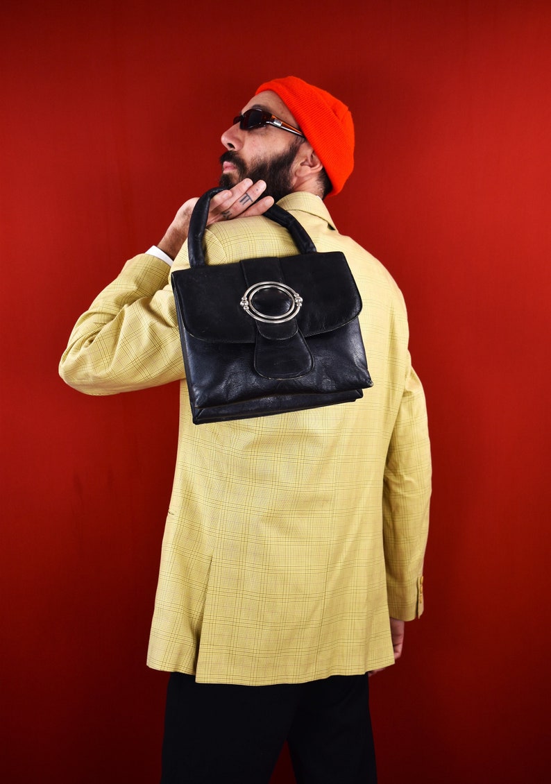 May include: Black leather handbag with a silver-tone clasp. The bag has a structured shape and a top handle. The person is wearing a yellow plaid blazer and an orange beanie.