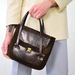 May include: A brown handbag with a snakeskin pattern and suede accents. The bag has a rectangular shape with a gold-colored clasp and two handles. The bag is held by a person wearing a cream-colored blazer.
