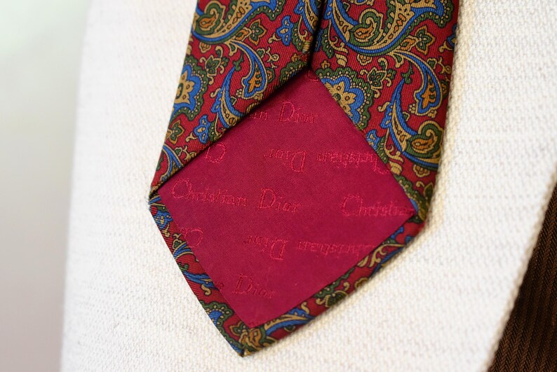 Pu&ograve; includere: Una cravatta di seta con motivo paisley rosso e verde con una fodera rossa su cui &egrave; scritto "Christian Dior" in lettere bianche.