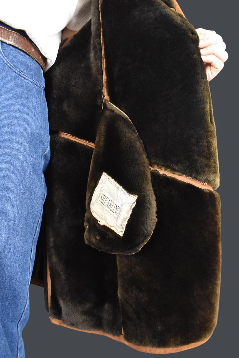 May include: A brown shearling jacket with a brown leather trim. The jacket is lined with a soft, black shearling fabric. The label on the jacket reads "Shearling".
