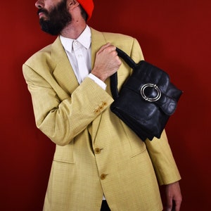 May include: A yellow plaid blazer with a white shirt and black pants. The person is holding a black leather handbag with a silver buckle. The background is a solid red.