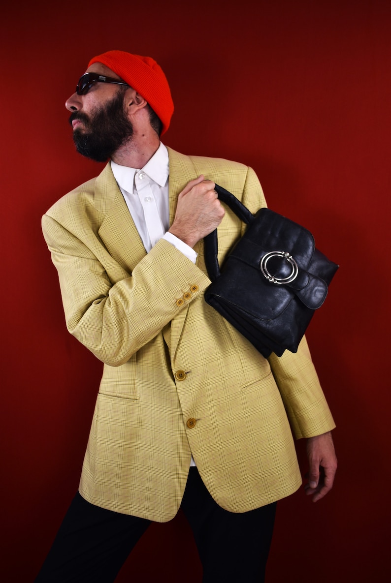 May include: A yellow plaid blazer with a white shirt and black pants. The person is holding a black leather handbag with a silver buckle. The background is a solid red.