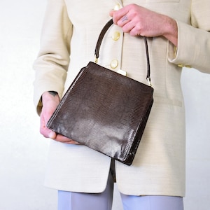 Könnte beinhalten: Eine braune, strukturierte Handtasche mit rechteckiger Form und einem oberen Griff. Die Tasche hat einen Schnallenverschluss und ein subtiles, reptilienartiges Muster. Der Griff ist mit goldfarbener Hardware befestigt.