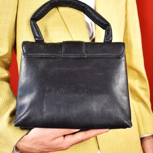 May include: Black leather handbag with a top handle and a rectangular shape. The bag features a flap closure with a buckle. The handle is padded for comfort. The bag is held against a yellow plaid blazer.