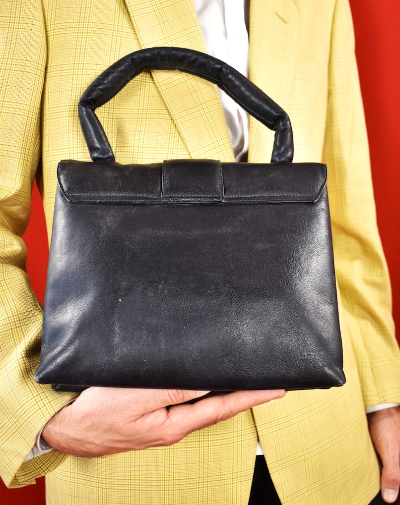 May include: Black leather handbag with a top handle and a rectangular shape. The bag features a flap closure with a buckle. The handle is padded for comfort. The bag is held against a yellow plaid blazer.