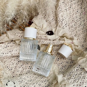 Puede incluir: Dos frascos de perfume de vidrio transparente con tapas blancas y doradas. Los frascos tienen un diseño acanalado y se exhiben sobre una tela crema texturizada. Una tapa está quitada, revelando la boquilla dorada.