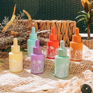 Könnte beinhalten: Sechs Glas-Tropfflaschen mit floralen Designs in verschiedenen Pastellfarben, darunter Gelb, Grün, Blau und Lila, auf einer weißen Häkel-Oberfläche angeordnet.