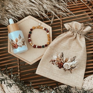 Könnte beinhalten: Ein Set aus drei Artikeln: eine kleine Flasche ätherisches Öl mit floralem Design, ein brauner Jutesack mit Kordelzug und floralem Design sowie ein Perlenarmband mit roten, gelben und grünen Perlen.