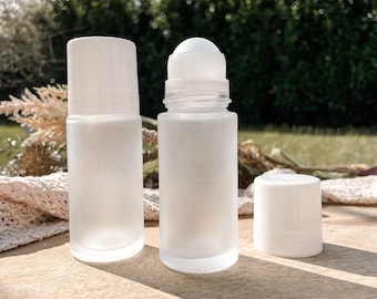 May include: Two frosted glass roll-on bottles, one with a cap and one without, and a separate white cap. The bottles are cylindrical with a rounded top for the rollerball. The background is a wooden surface with greenery.