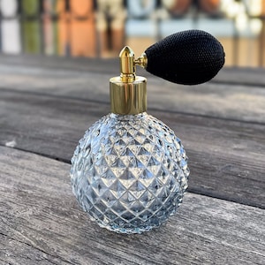 Könnte beinhalten: Eine durchsichtige Glas-Parfümflasche mit goldenem Verschluss und schwarzem Zerstäuber. Die Flasche hat ein strukturiertes, rautenförmiges Muster.