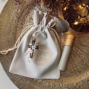 Könnte beinhalten: Ein beiger Kordelzugbeutel mit Kreuzdesign und den Worten "DU BIST... Geliebt" liegt auf einem goldfarbenen, strukturierten Teller. Daneben befindet sich eine passende graue Flasche mit Holzdeckel. Trockenblumen und Lichter im Hintergrund.