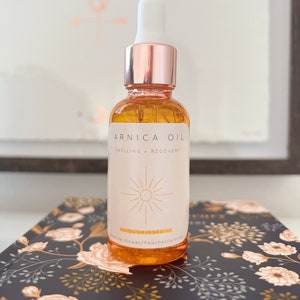 Puede incluir: Una botella de vidrio de aceite de árnica con una tapa y un gotero de oro rosa. La etiqueta es blanca con texto negro que dice "Arnica Oil, Swelling, Recovery". La botella está sobre una superficie con estampado floral blanco y negro. La etiqueta también presenta un gráfico de sol y el texto "Sunkissed, Jasmine Flower/Peaches/Orange".