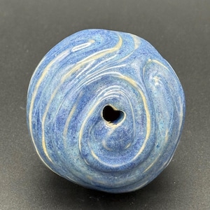 May include: A blue ceramic decorative object with a heart-shaped hole. The object has a swirled pattern in shades of blue and cream. The ceramic piece is round and has a glossy finish.