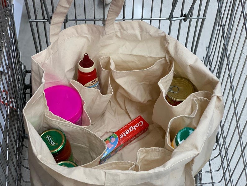 May include: A beige canvas tote bag with multiple compartments, filled with various grocery items, including a pink container, a red bottle of ketchup, a green jar labeled "Dolmio", a tube of Colgate toothpaste, and a can of food.