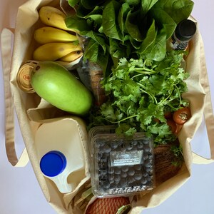 May include: A white canvas tote bag filled with groceries, including bananas, a green mango, spinach, cilantro, blueberries, a carton of milk, and a bottle of dark liquid.