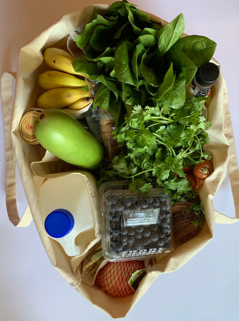 May include: A white canvas tote bag filled with groceries, including bananas, a green mango, spinach, cilantro, blueberries, a carton of milk, and a bottle of dark liquid.