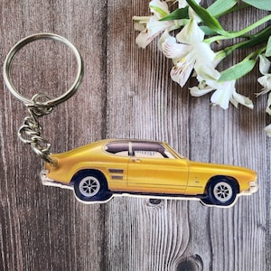 Może przedstawiać: Złoty brelok do kluczy z samochodem ze srebrnym kółkiem i łańcuszkiem. Samochód to klasyczny design coupe z czarnymi oponami i szczegółową ilustracją. Brelok znajduje się na drewnianym tle.
