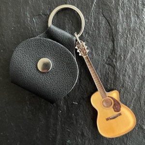 Può includere: Un porta plettri in pelle nera con chiusura a bottone automatico argentato e portachiavi argentato. Un ciondolo a forma di chitarra acustica in miniatura è attaccato al portachiavi. Il ciondolo della chitarra è marrone chiaro con una buca scura.