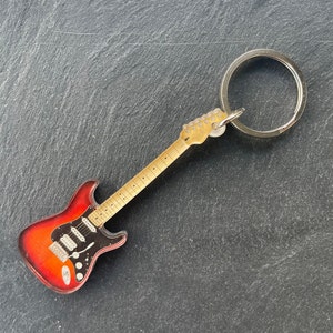 Può includere: Un portachiavi a forma di chitarra elettrica rossa e nera con un anello portachiavi argentato. La chitarra ha una finitura in legno e un battipenna nero.
