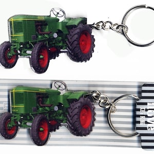 Könnte beinhalten: Grüner Traktor-Schlüsselanhänger mit einem silbernen Schlüsselring. Der Traktor hat rote Räder und befindet sich auf einem gestreiften Hintergrund mit dem aufgedruckten Wort "love".
