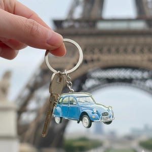 Puede incluir: Un llavero de coche azul vintage con un llavero plateado y una llave. El llavero presenta un coche azul en miniatura con ruedas blancas. La Torre Eiffel es visible en el fondo.