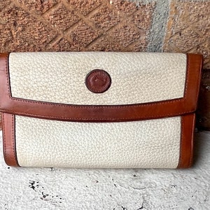 May include: A cream-colored clutch with brown leather trim and a flap closure. The clutch features a textured surface and a circular emblem on the front. The edges and flap are trimmed with brown leather.