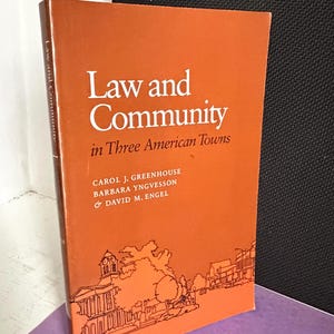 May include: A hardcover book titled Law and Community in Three American Towns by Carol J. Greenhouse, Barbara Yngvesson, and David M. Engel. The cover is a solid burnt orange color with white text and a dark orange line drawing of a town square.