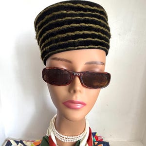 May include: A black and olive green striped pillbox hat worn with brown tortoiseshell sunglasses featuring the letters DG on the bridge and a triple-strand white pearl necklace paired with a green, red, and white patterned silk scarf.
